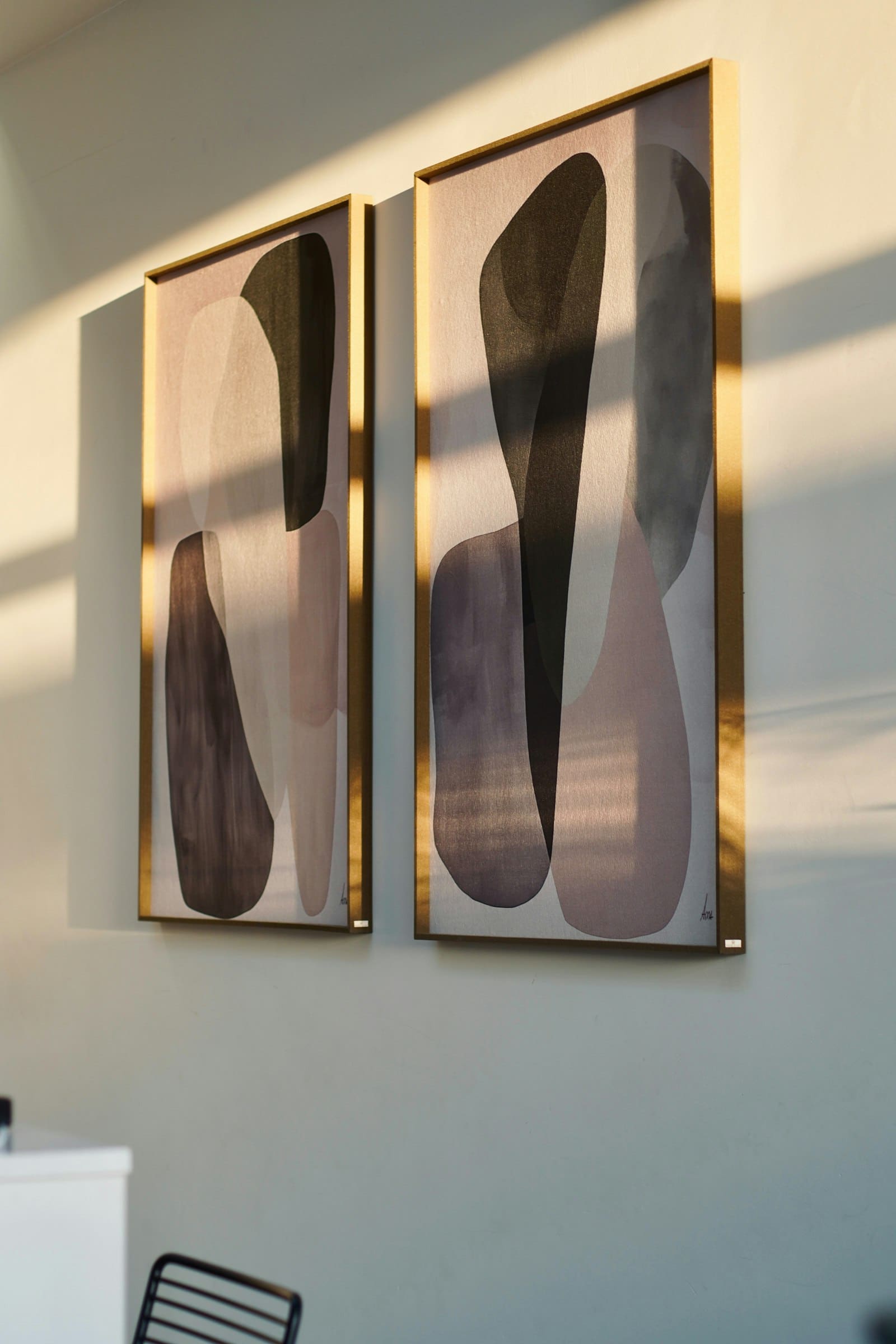 Two abstract prints in slim brass frames on a sunlit warm beige wall